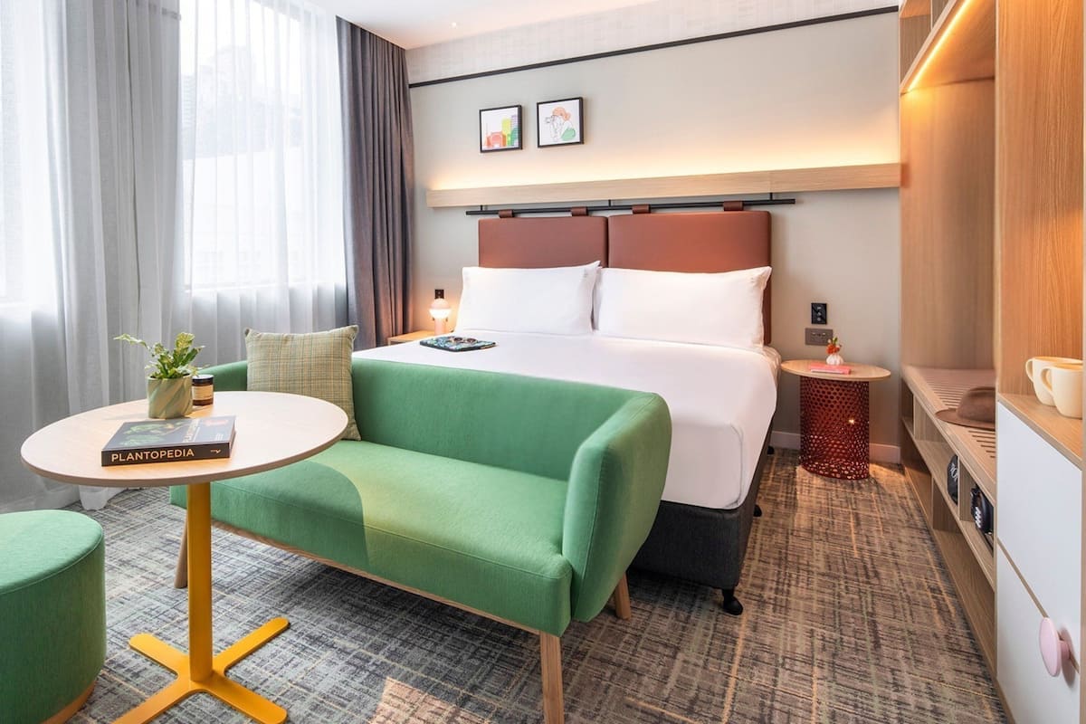 Une chambre d’hôtel avec un lit, un canapé vert, une petite table ronde et une table de chevet avec une lampe.
