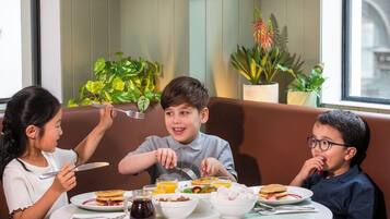A dining table with plates of food, a bottle of syrup, and a glass of orange juice. There are three people seated around the table.