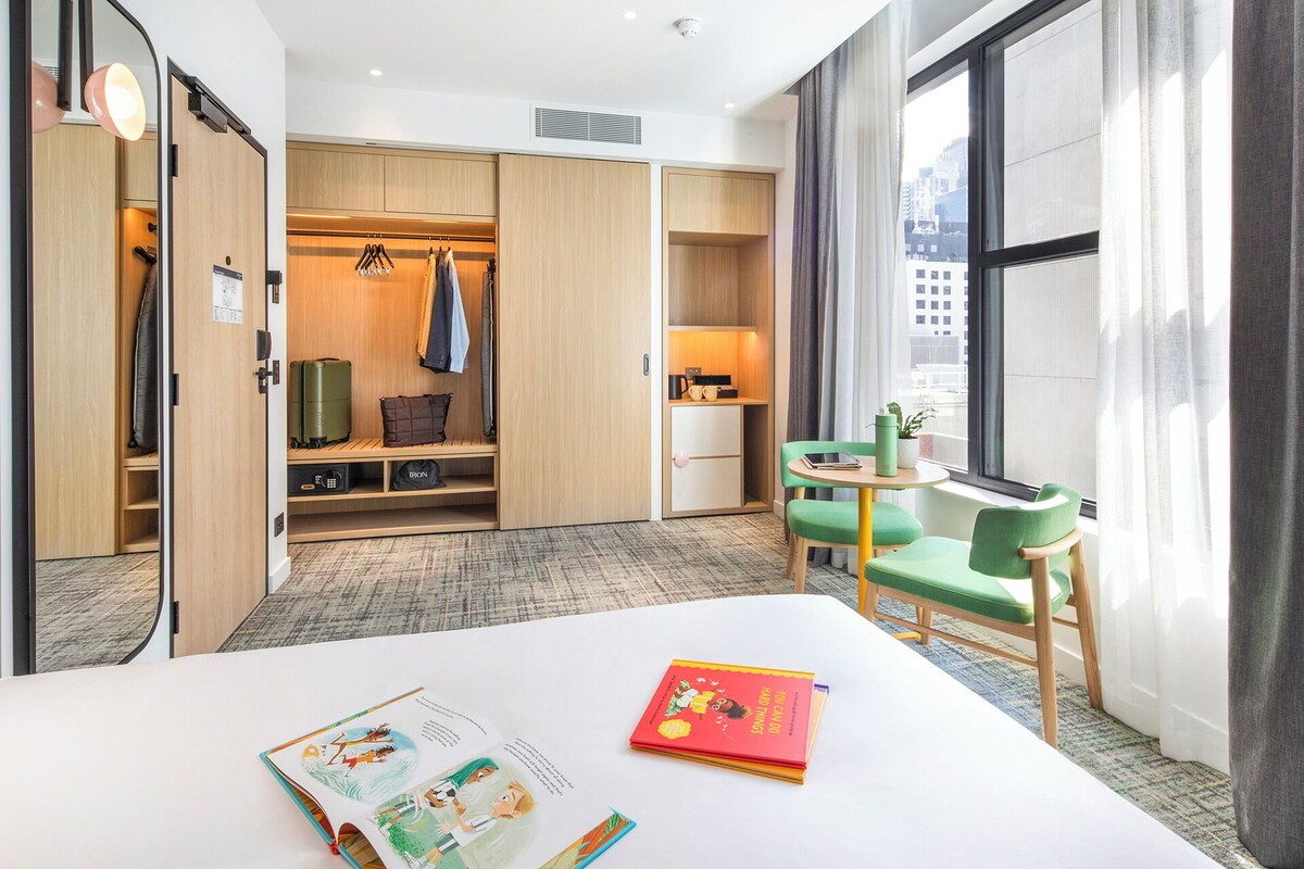Een hotelkamer met een bed, een bureau met stoel, een kledingkast, een kleine tafel met een plant en een raam met uitzicht op de stad.