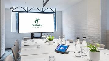 Una sala conferenze con un grande schermo che mostra il logo di Holiday Inn, un tavolo bianco con bottiglie d'acqua, un laptop e una pianta in vaso.