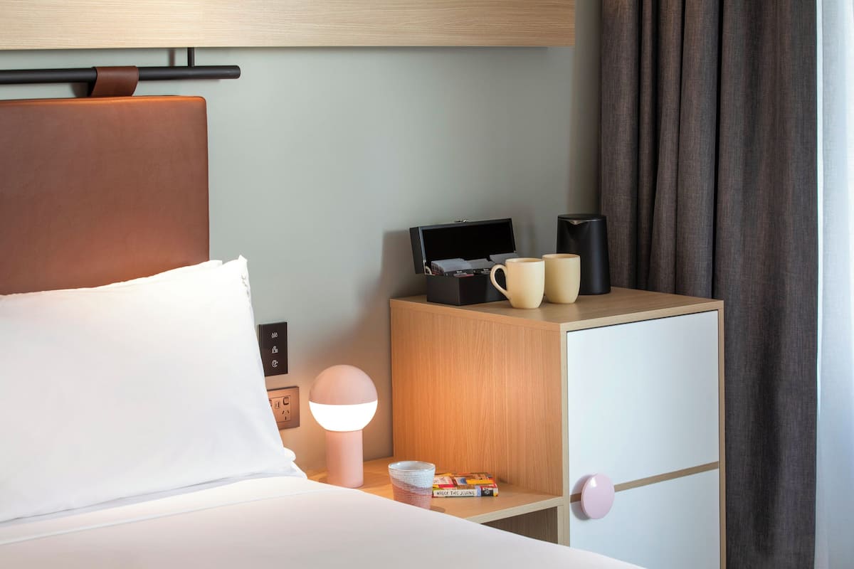 Une chambre d’hôtel bien rangée, avec un lit, une table de chevet et une petite armoire en bois contenant une lampe, une machine à café et une tasse.