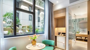 Een moderne hotelkamer met een groot raam, een ronde tafel met twee groene stoelen, een kleine vaas met bloemen en een houten kast.