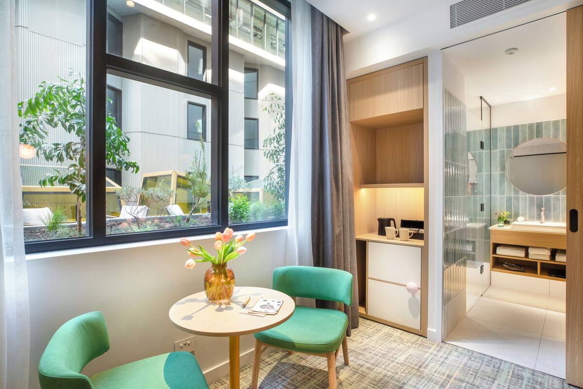 Une chambre d’hôtel moderne avec une grande fenêtre, une table ronde et deux chaises vertes, un petit vase avec des fleurs et un meuble en bois.