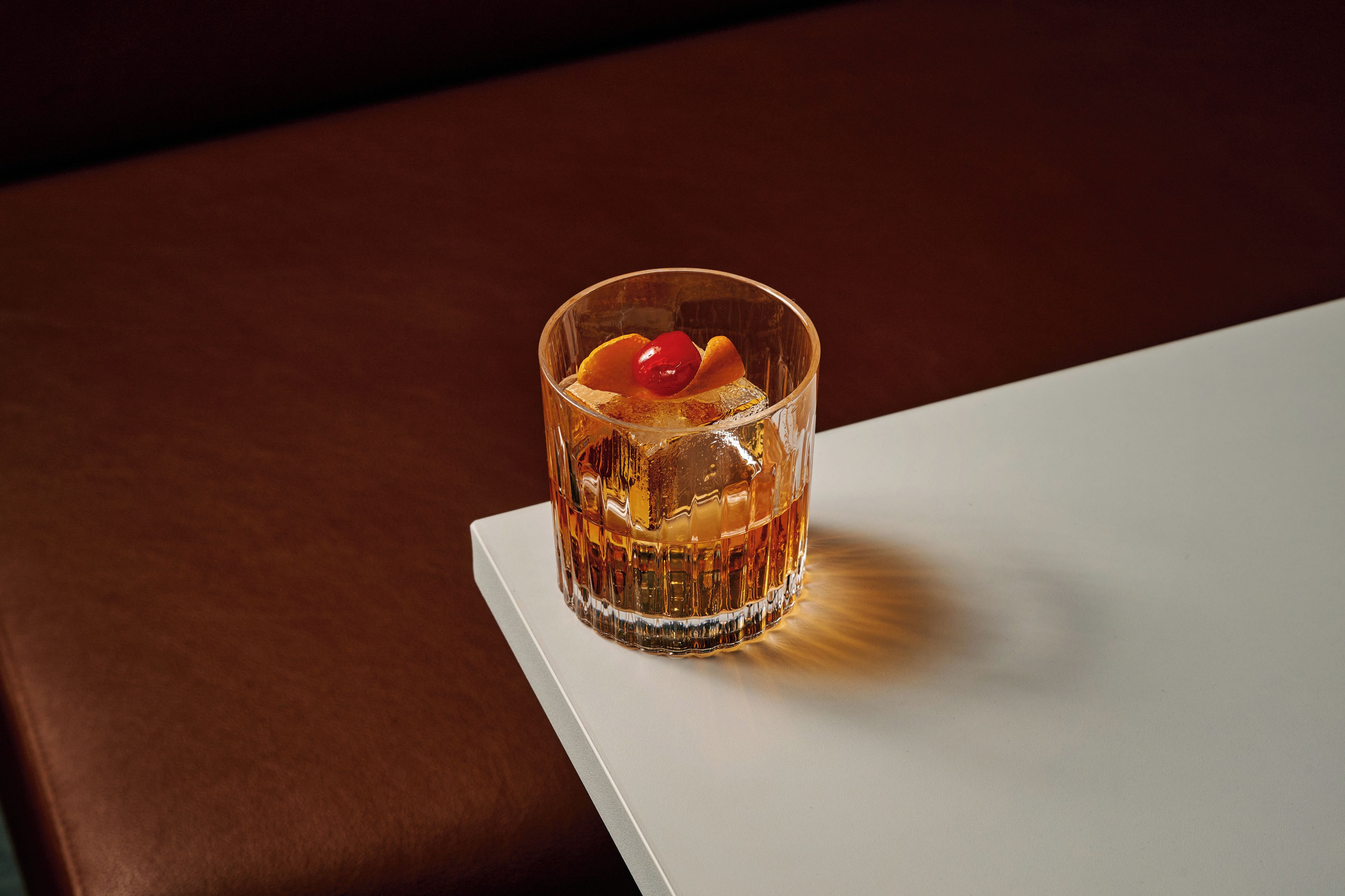 Ein Glas mit bernsteinfarbiger Flüssigkeit, Eiswürfeln und einer roten Dekoration auf weißer Oberfläche.