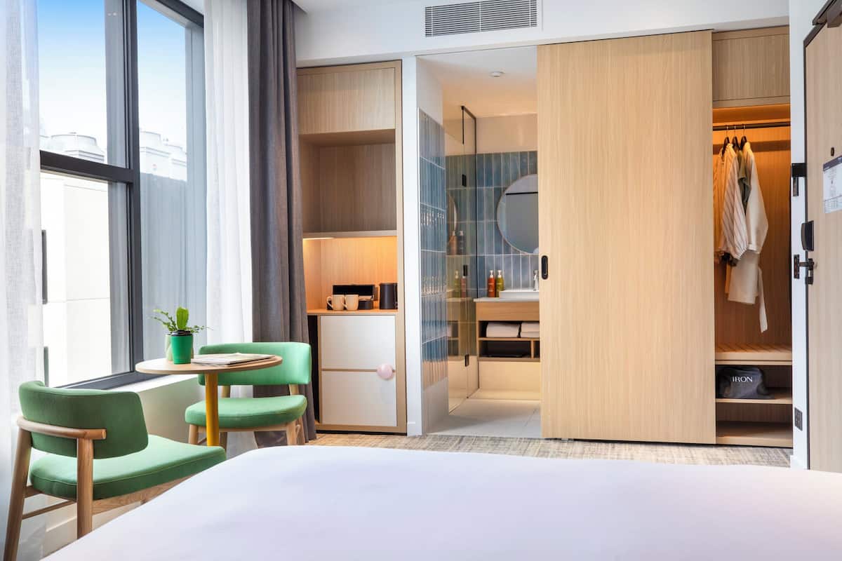 Une chambre d’hôtel comprenant un lit, un bureau avec une chaise, une petite table, une plante en pot, une armoire et une salle de bain avec un miroir.