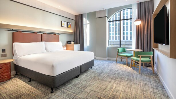 Une chambre d’hôtel avec un grand lit, une télévision, une petite table, deux chaises et une fenêtre avec des rideaux.