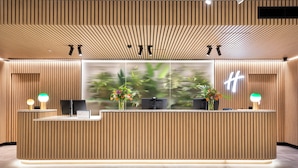 Een receptiebalie met verticale houten lambrisering, twee computermonitors en een groot raam met een onscherp uitzicht op groen.