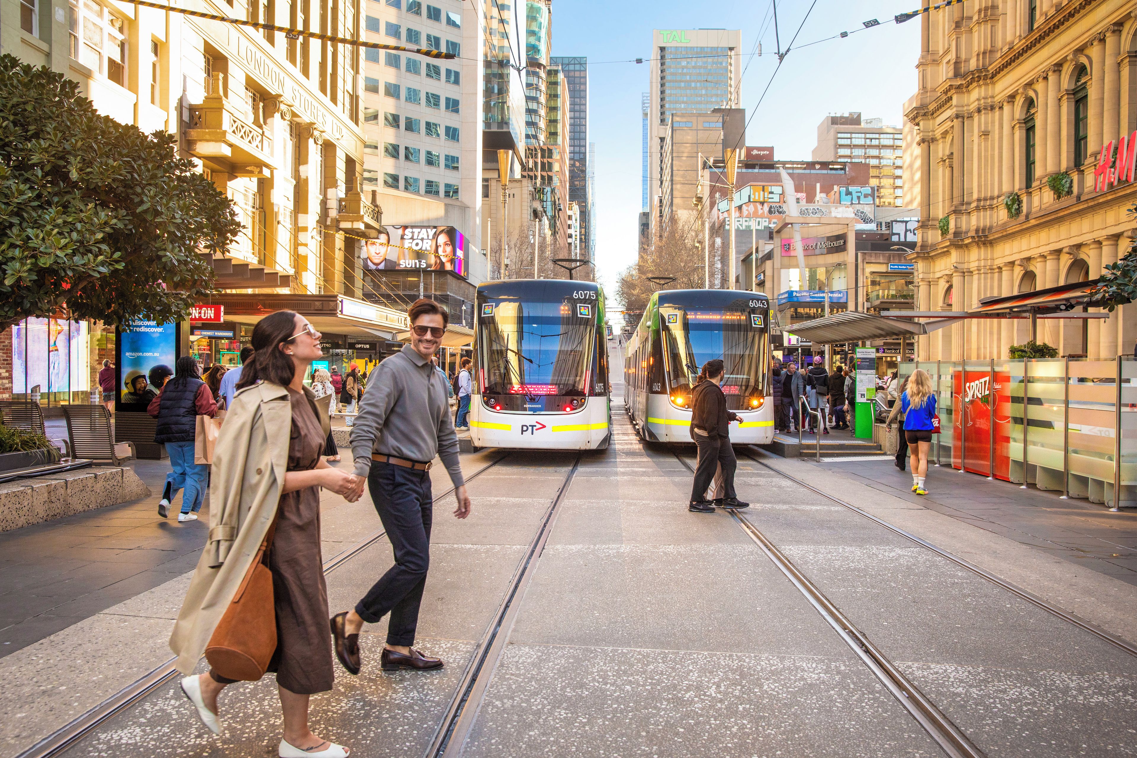 A tram station with two trams, a pedestrian walkway, and urban buildings.