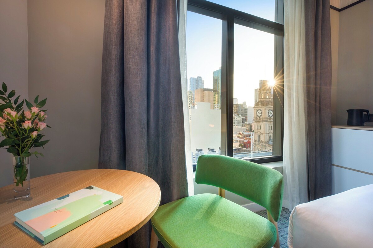 Een hotelkamer met uitzicht op een stadsbeeld, een groene stoel, een houten tafel met een boek en een vaas met bloemen.