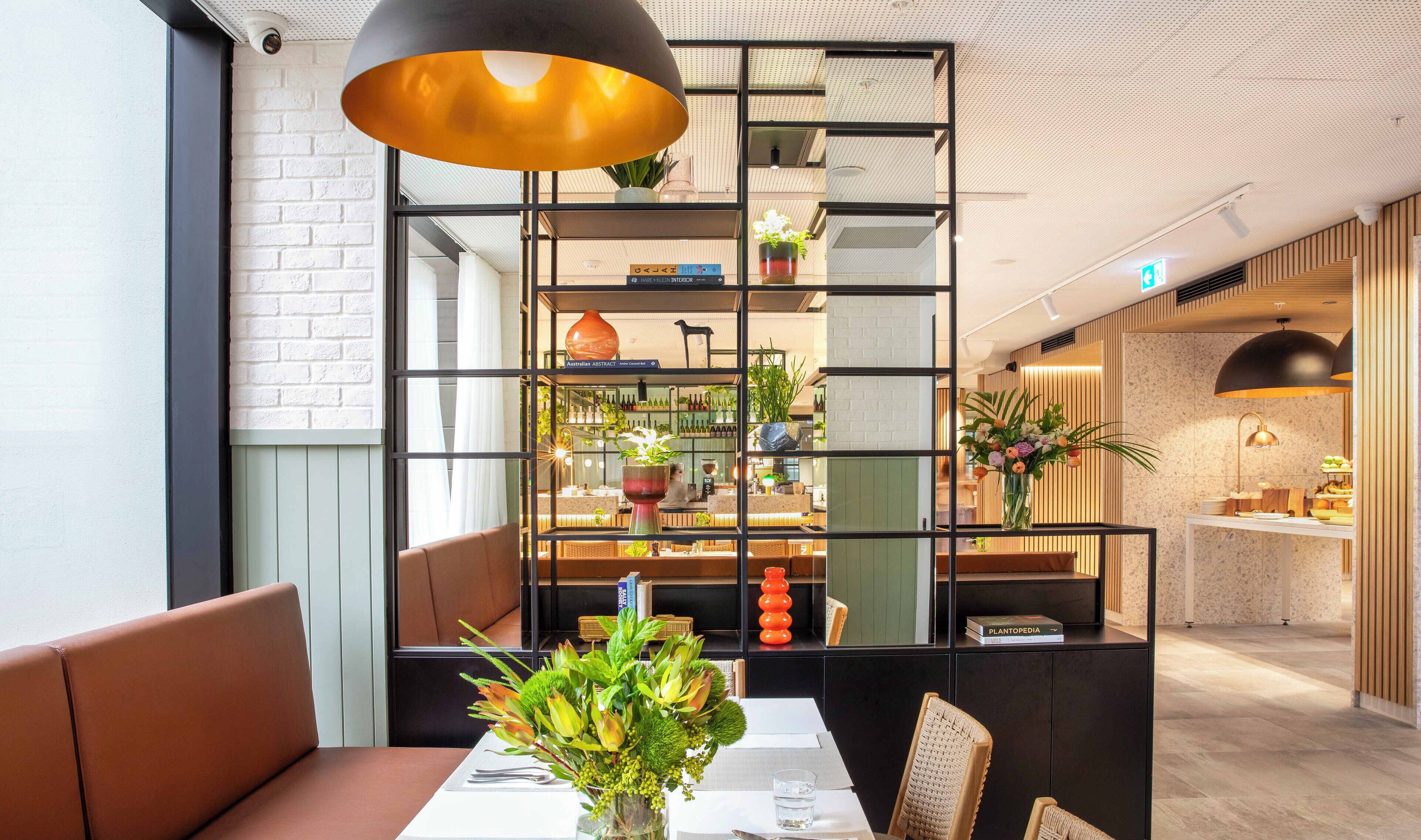 Ein modernes Interieur mit einer Essecke, komplett gedecktem Tisch für zwei Personen, einem schwarzen Metallregal mit Pflanzen und einer warmen Pendelleuchte.
