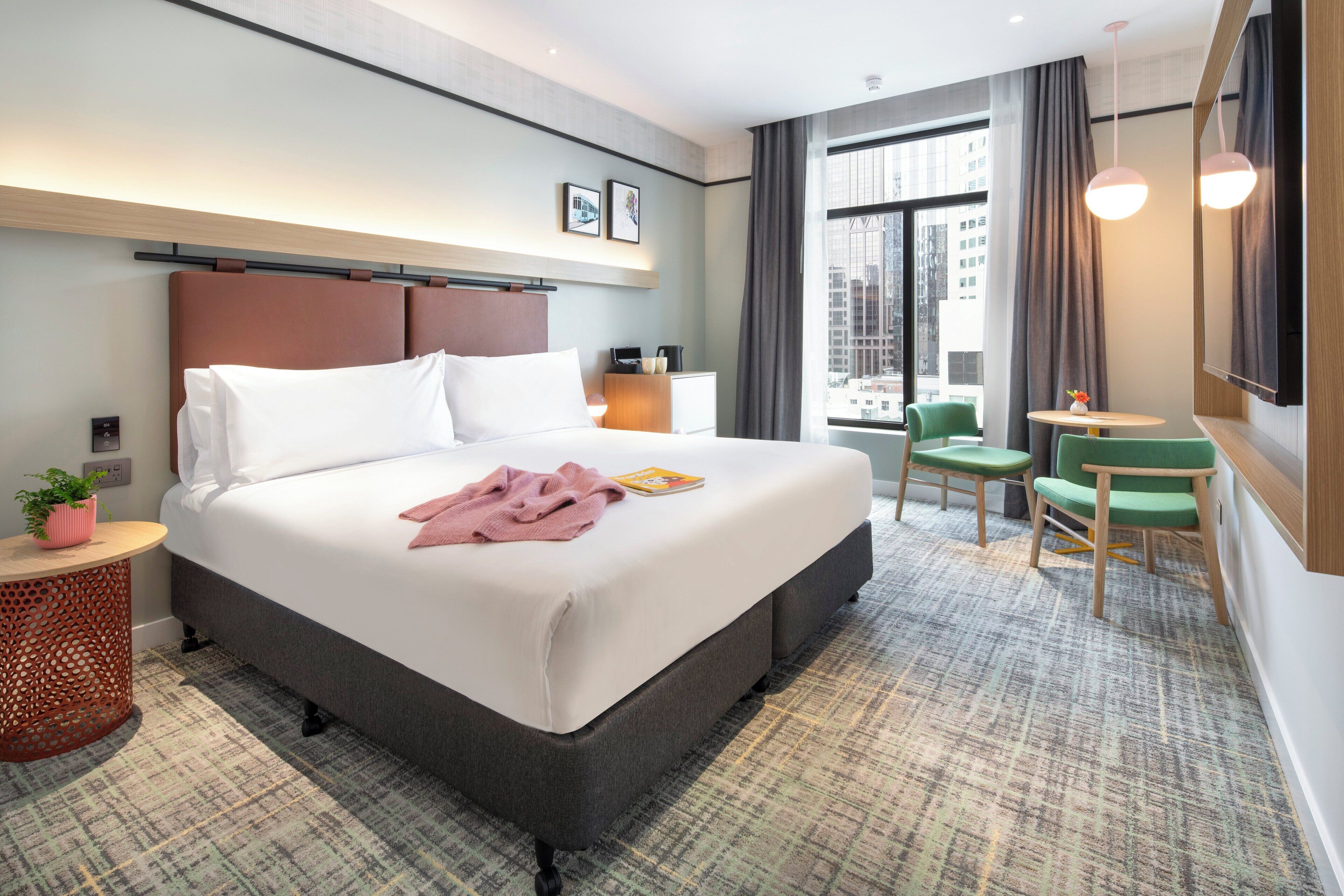 A modern hotel room with a large bed, a small table, two chairs, a TV, and a large window with a city view.