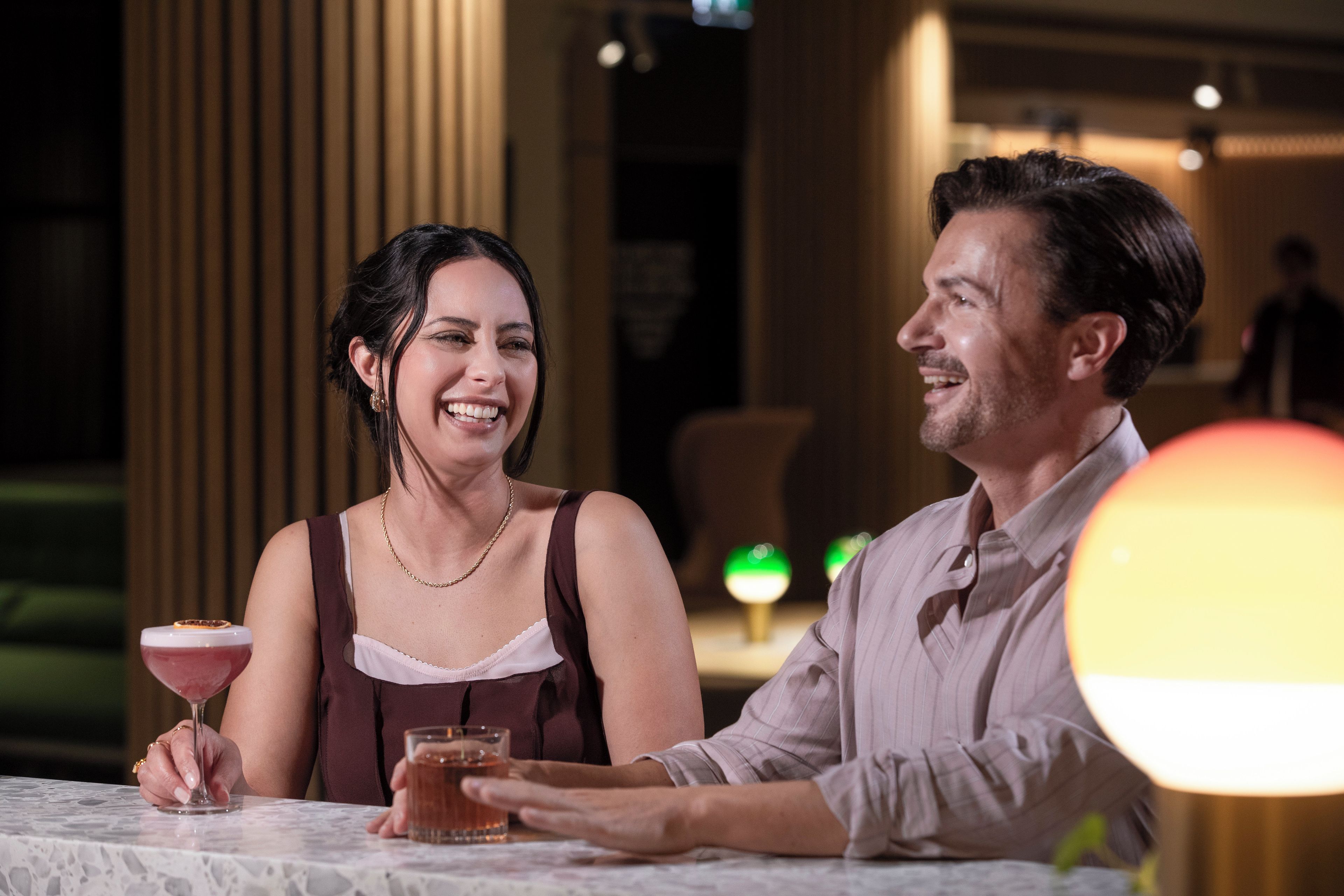 Una pareja sentada en una mesa con bebidas, en un ambiente interior con poca luz.
