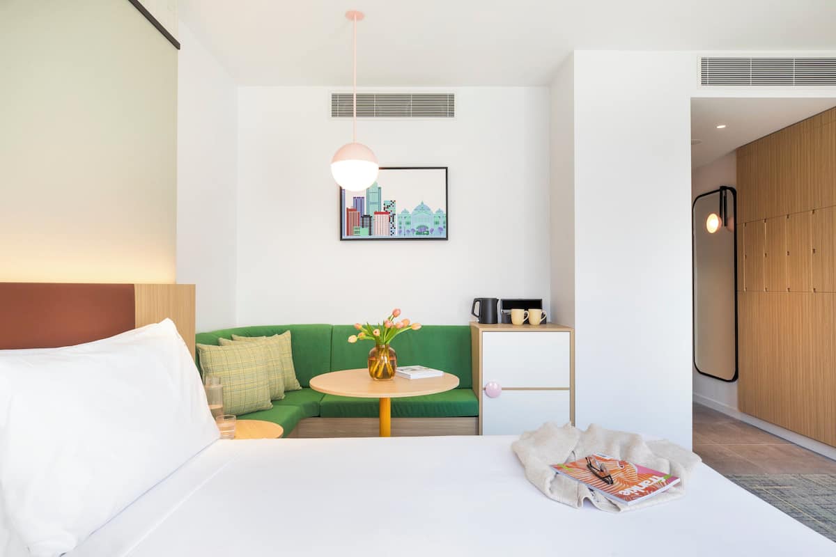 Une chambre d’hôtel moderne avec un lit, un canapé vert, une petite table ronde ornée d’un vase de fleurs, un tableau encadré représentant un paysage urbain et une armoire en bois.