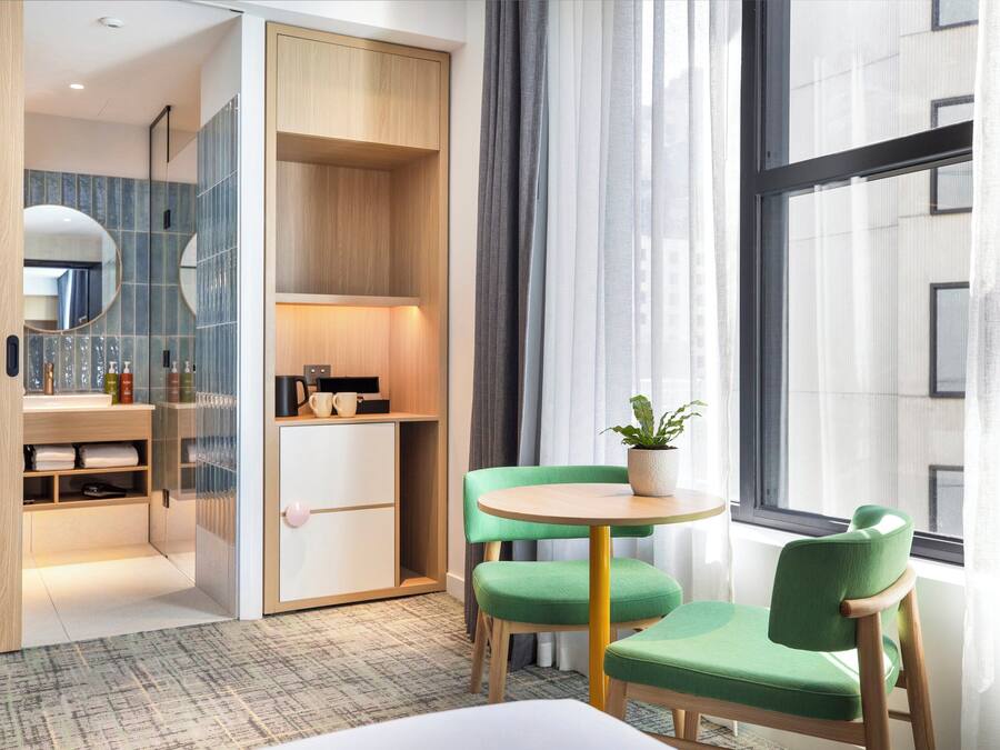 Habitación de hotel moderna con un gran ventanal, una silla verde, una mesita con una planta y un armario empotrado con espejo.