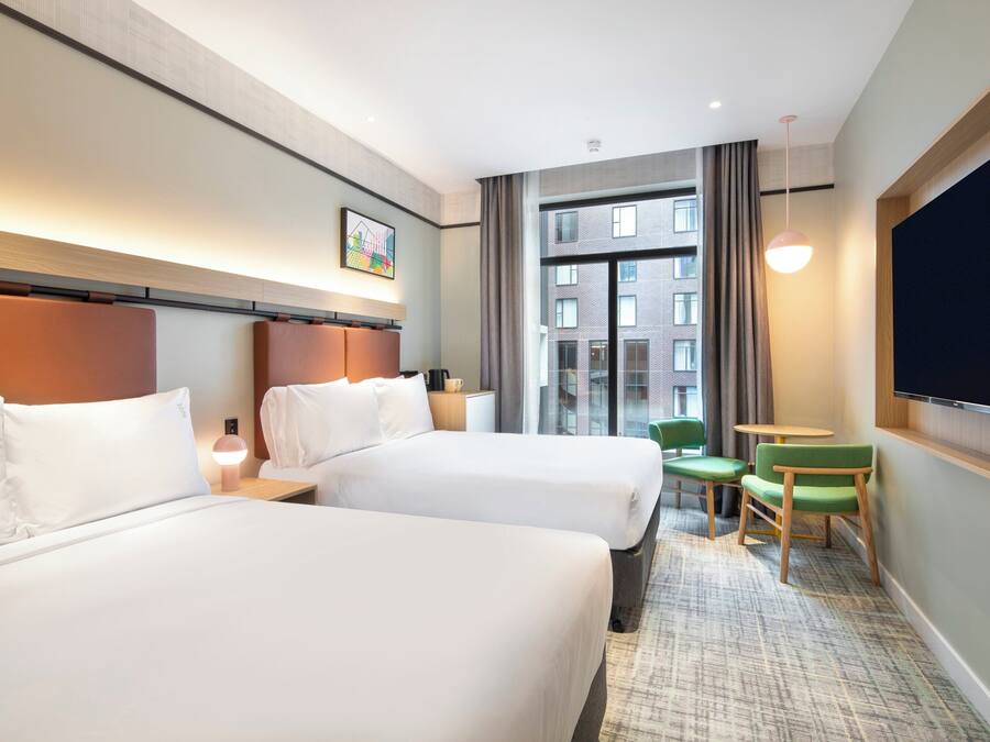Une chambre d’hôtel avec deux lits, une petite table, une chaise, un téléviseur et une grande fenêtre donnant sur des immeubles.