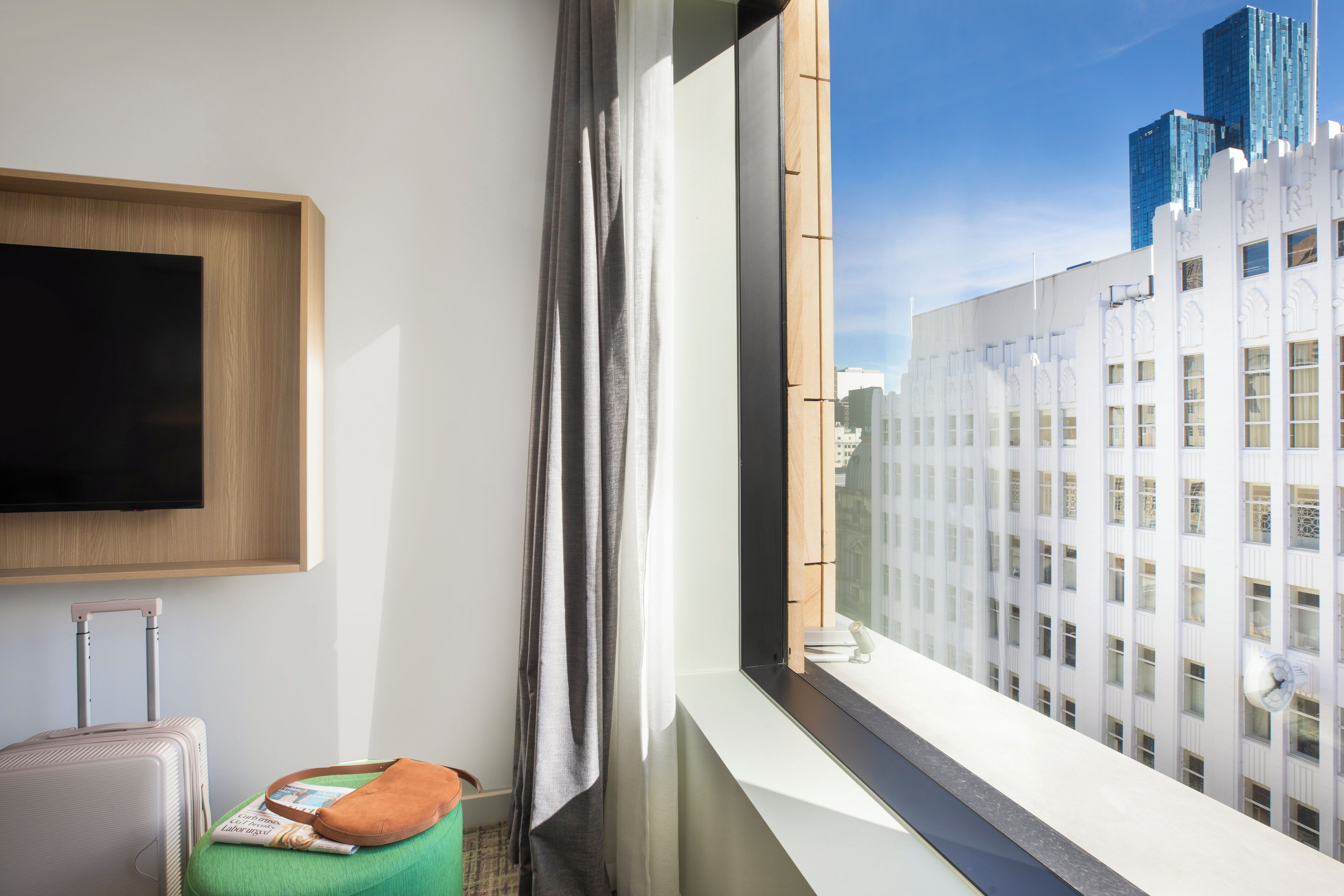 A hotel room with a large window, a flat-screen TV mounted on the wall, a suitcase, and a green cushion.