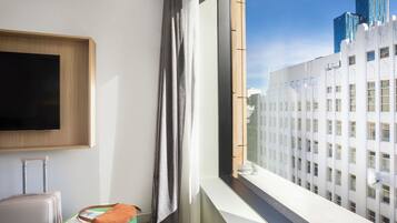 A hotel room with a large window, a flat-screen TV mounted on the wall, a suitcase, and a green cushion.