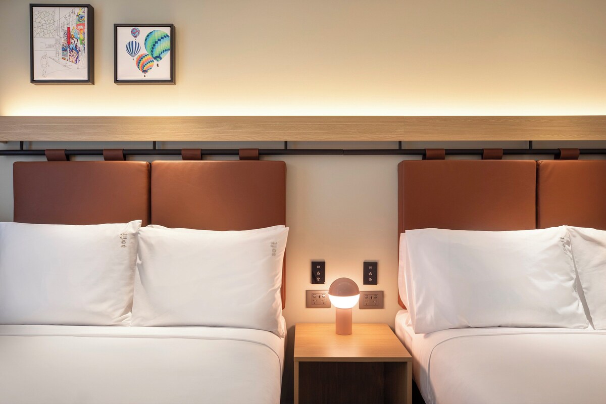 Een hotelkamer met twee bedden, een nachtkastje met een lamp en twee ingelijste kunstwerken aan de muur.