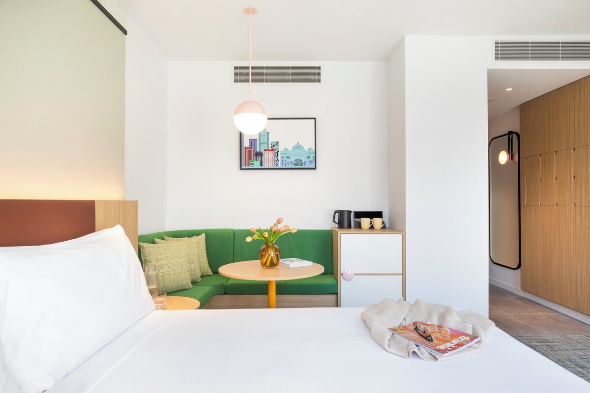 Een moderne hotelkamer met een bed, een groene bank, een klein rond tafeltje met een bloevas, een stadsgezicht in een lijst en een houten garderobe.