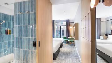 Quarto de hotel com área de chuveiro, duas camas, uma mesinha e vista para a cidade.