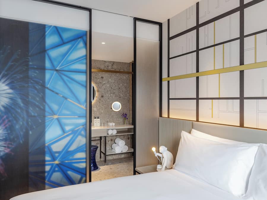 A hotel room with a bed, a bathroom with a mirror, and a wall with a geometric pattern.