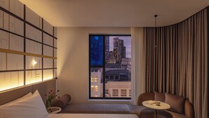 Habitación de hotel moderna con una cama grande, un área de descanso con mesa y vistas a la ciudad a través de un amplio ventanal.