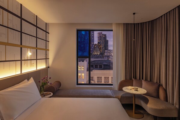 Habitación de hotel moderna con una cama grande, un área de descanso con mesa y vistas a la ciudad a través de un amplio ventanal.