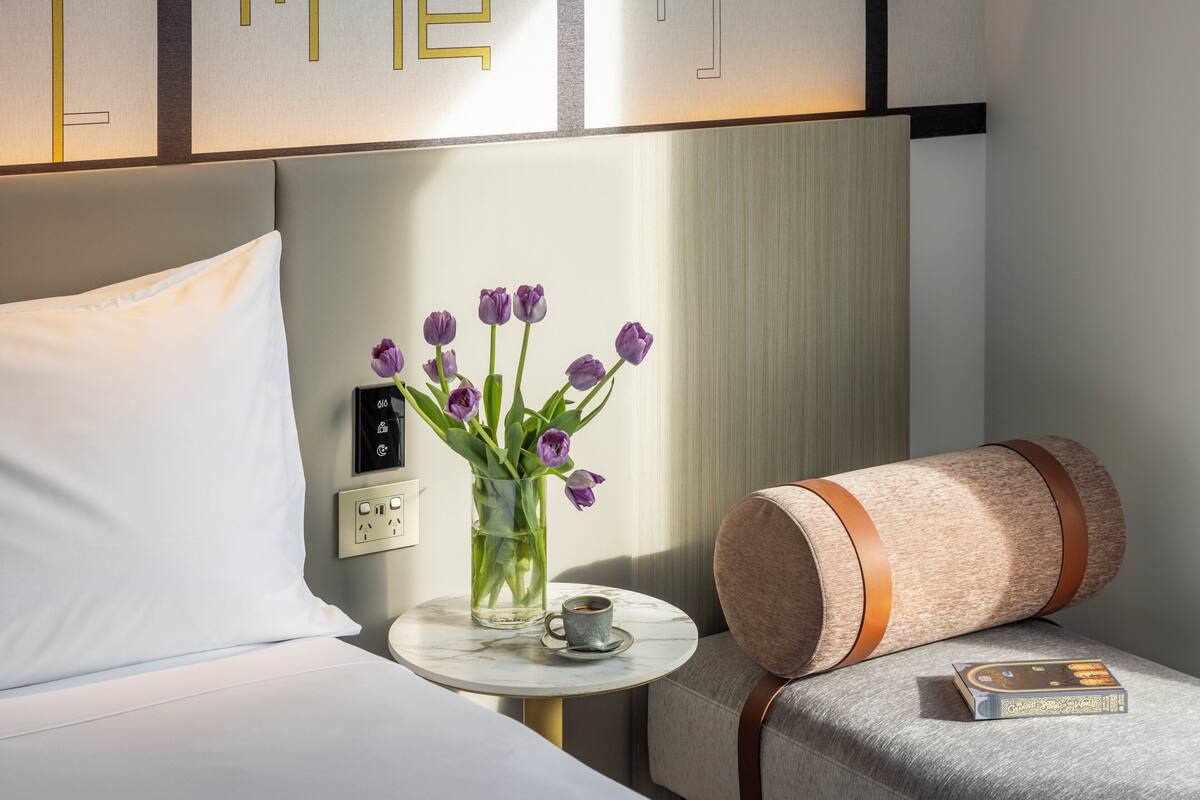 Ein Hotelzimmer mit einem Bett, einem Nachttisch mit einer Vase voller lila Blumen, einer Tasse und einem Buch.