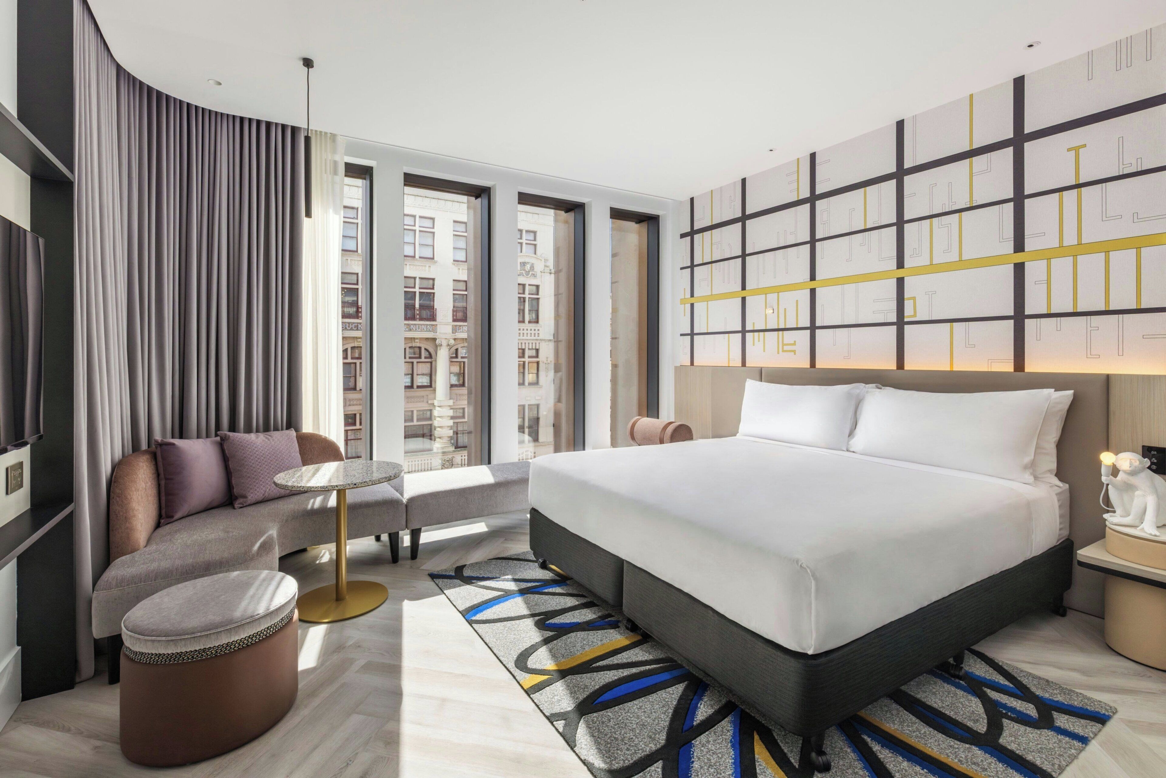 Quarto de hotel moderno com uma cama grande, área de estar com uma mesa e vista da cidade através de amplas janelas.