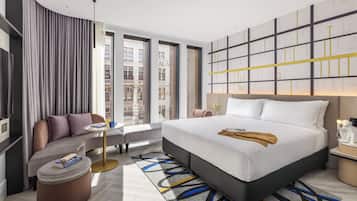 Ett modernt hotellrum med en stor säng, en soffa, en rund pall och utsikt över staden genom stora fönster.