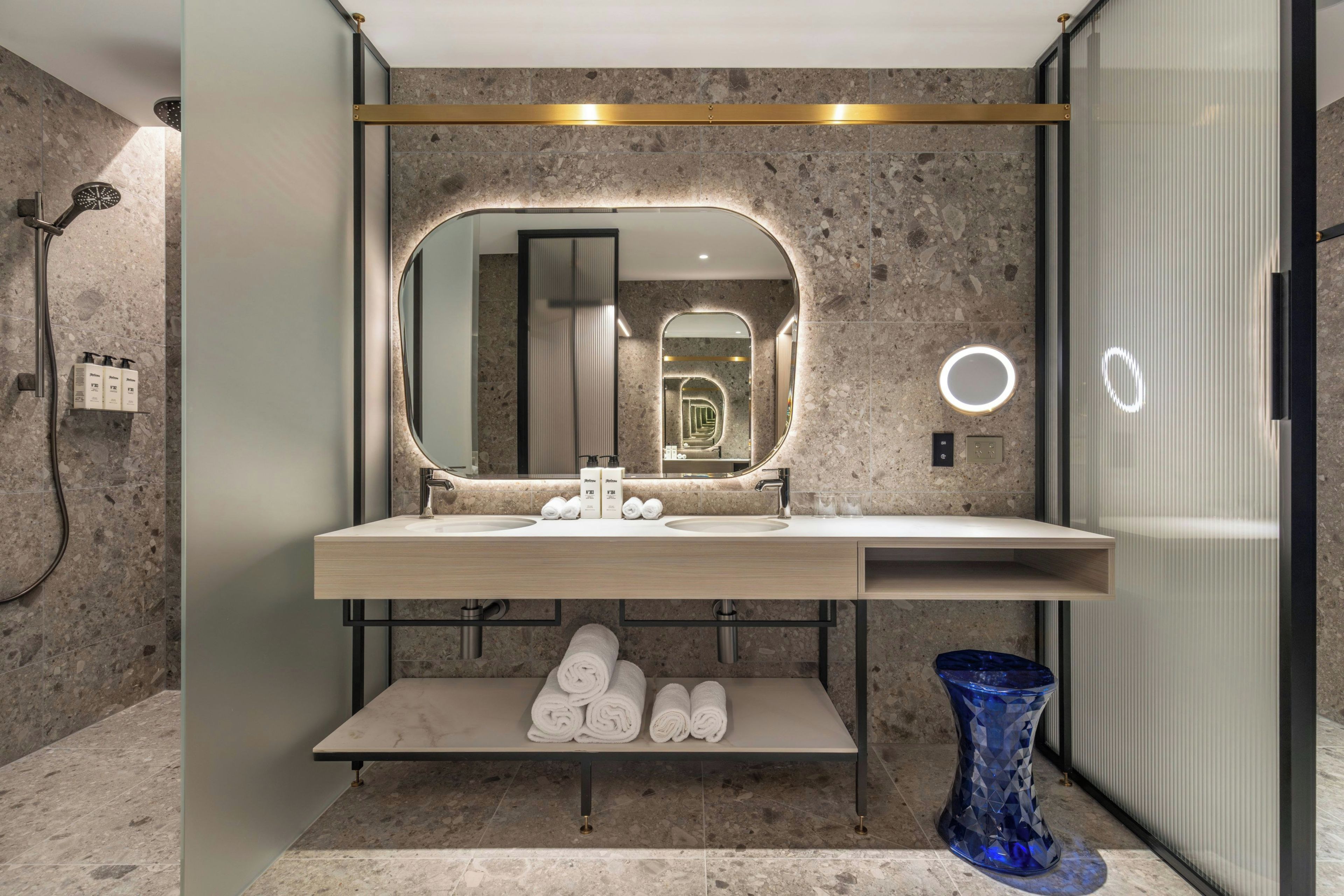 A modern bathroom with a marble floor, a large mirror with lights, a shower, and a blue vase.