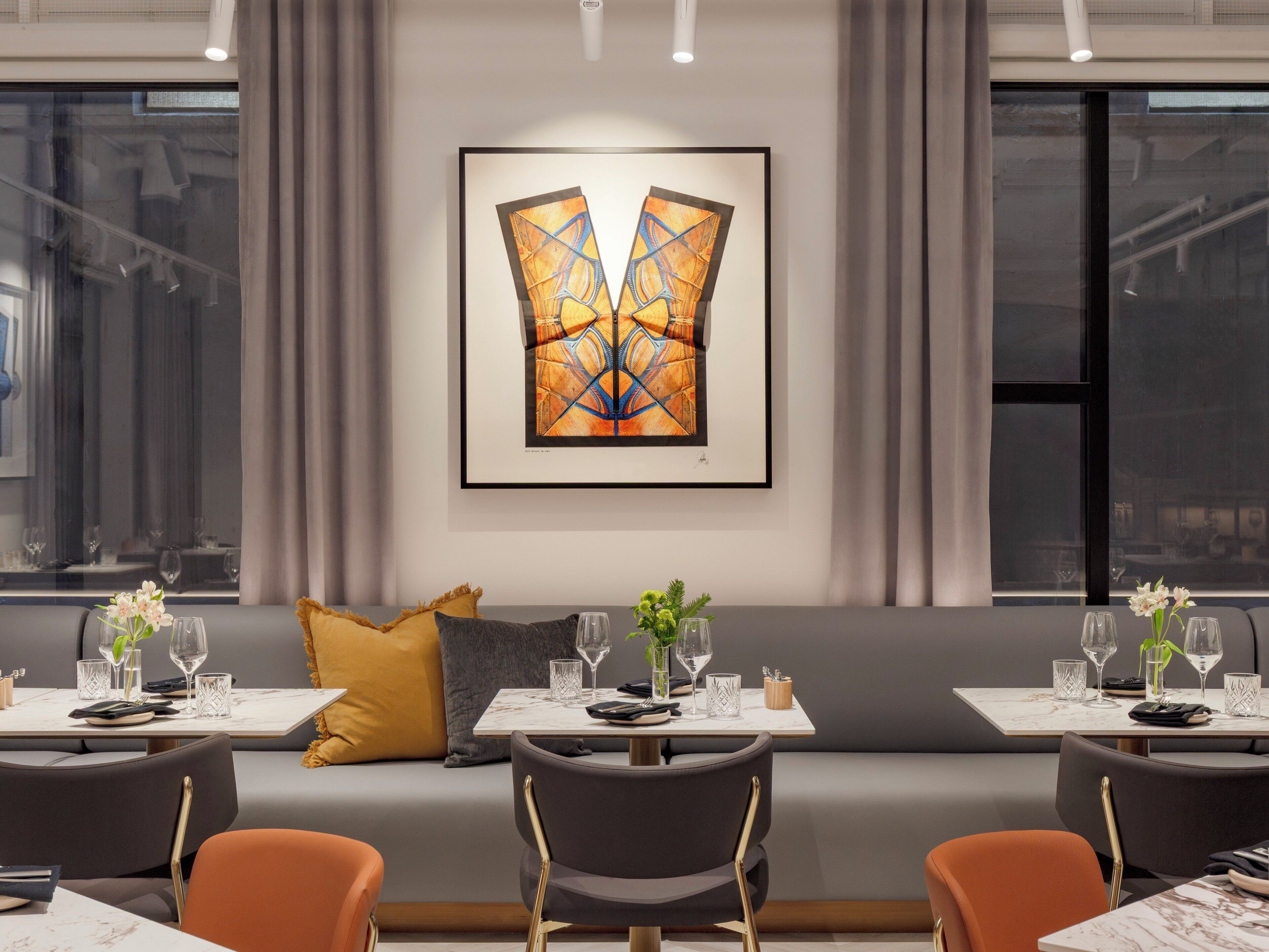 A modern dining area with a sofa and tables, a framed abstract painting on the wall, and a view of the city through large windows.