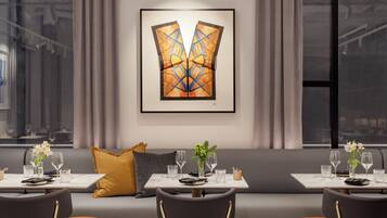A modern dining area with a sofa and tables, a framed abstract painting on the wall, and a view of the city through large windows.