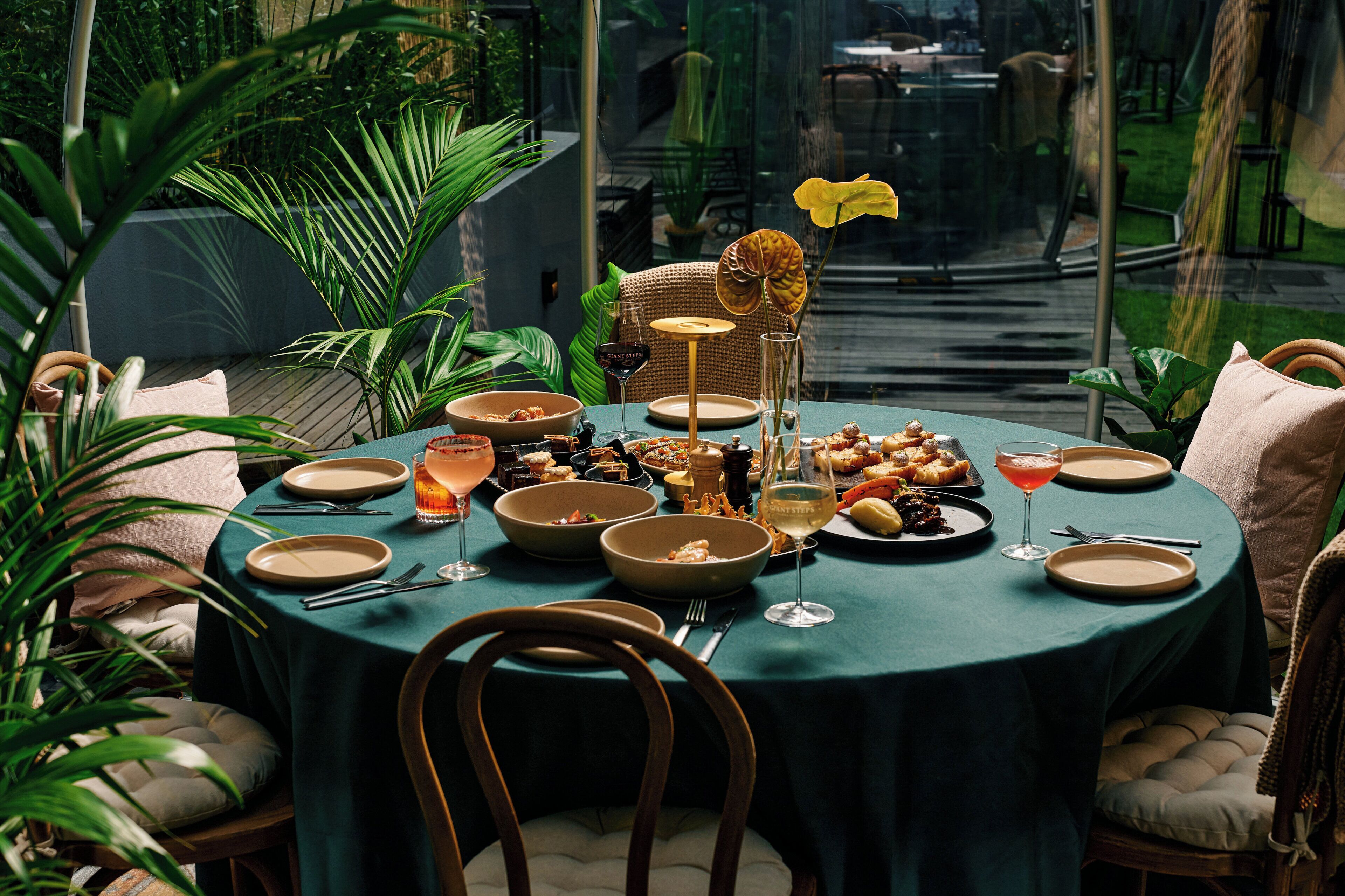 Un área de comedor al aire libre con una mesa redonda dispuesta para una comida, rodeada de sillas y plantas.
