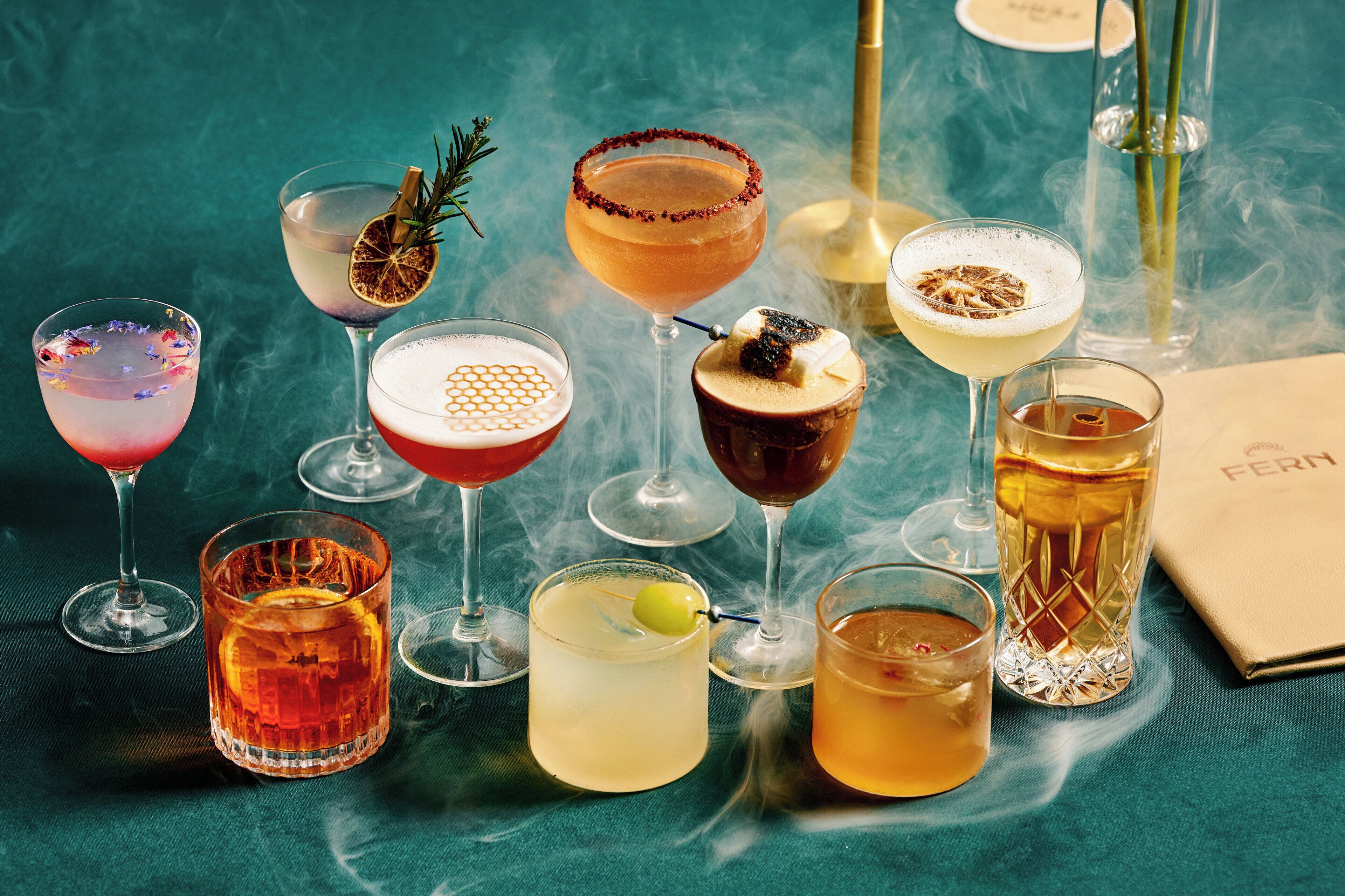 Diversas bebidas em copos diferentes sobre uma mesa com fundo verde-água.