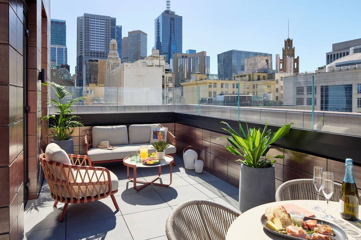Eine Dachterrasse mit Blick auf die Stadt, Korbmöbel, ein Esstisch mit einer Weinflasche und Topfpflanzen.