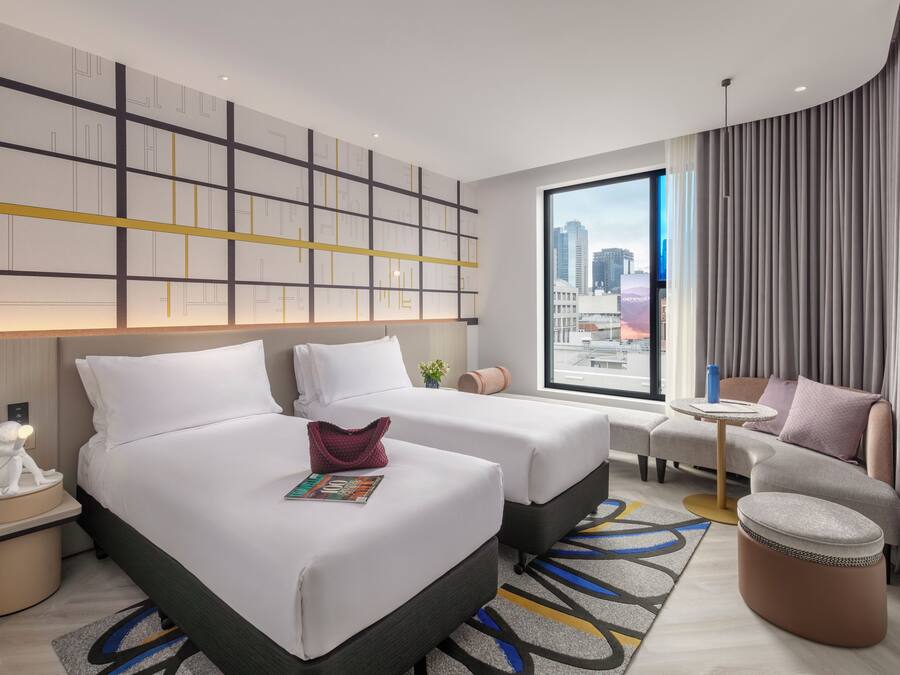 Ein modernes Hotelzimmer mit zwei Betten, einer Couch, einem kleinen Tisch und Blick auf die Stadt.