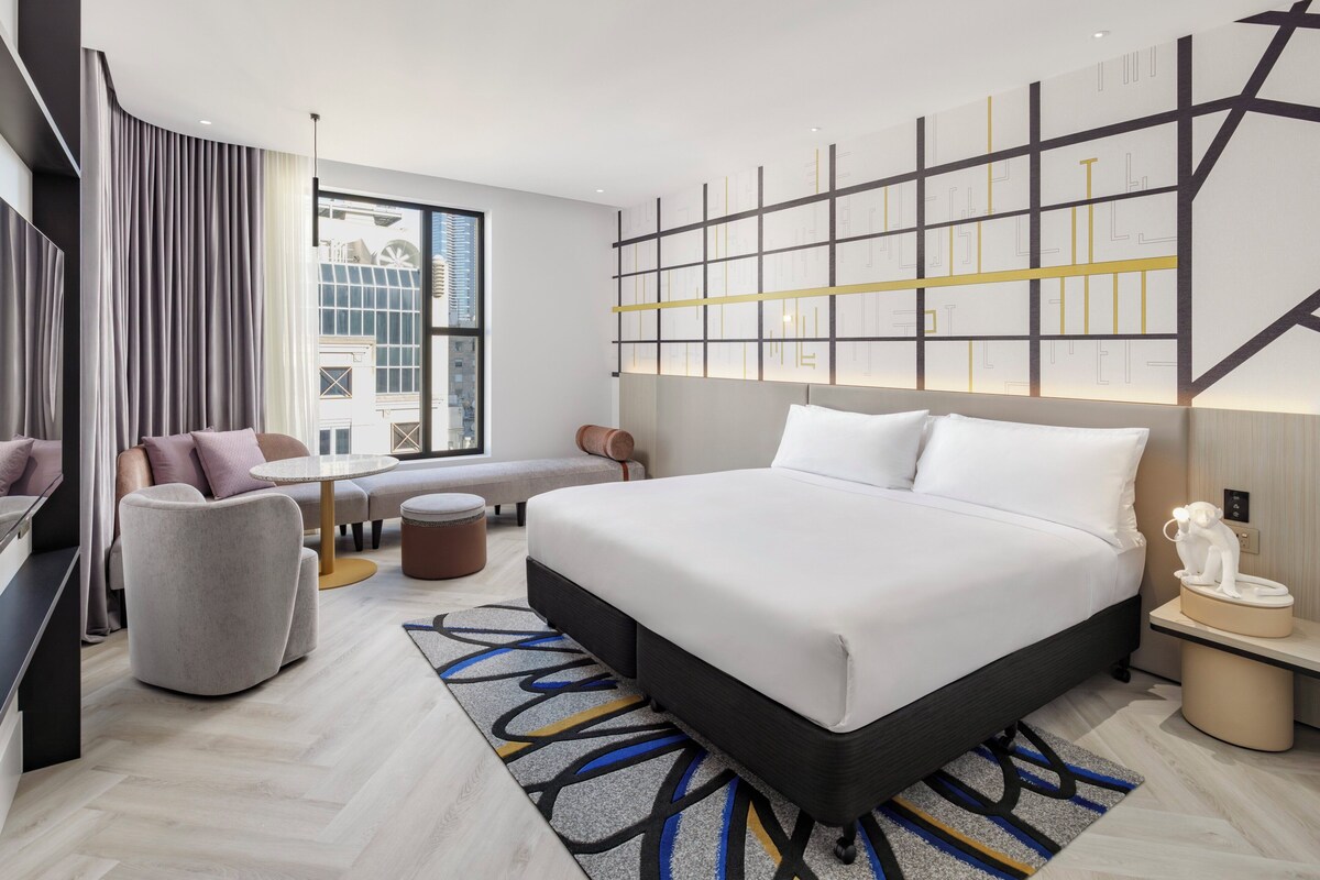 Ein modernes Hotelzimmer mit einem großen Bett, einer Sitzecke mit Sofa und Sesseln, einem kleinen Tisch und Blick auf die Stadt durch große Fenster.