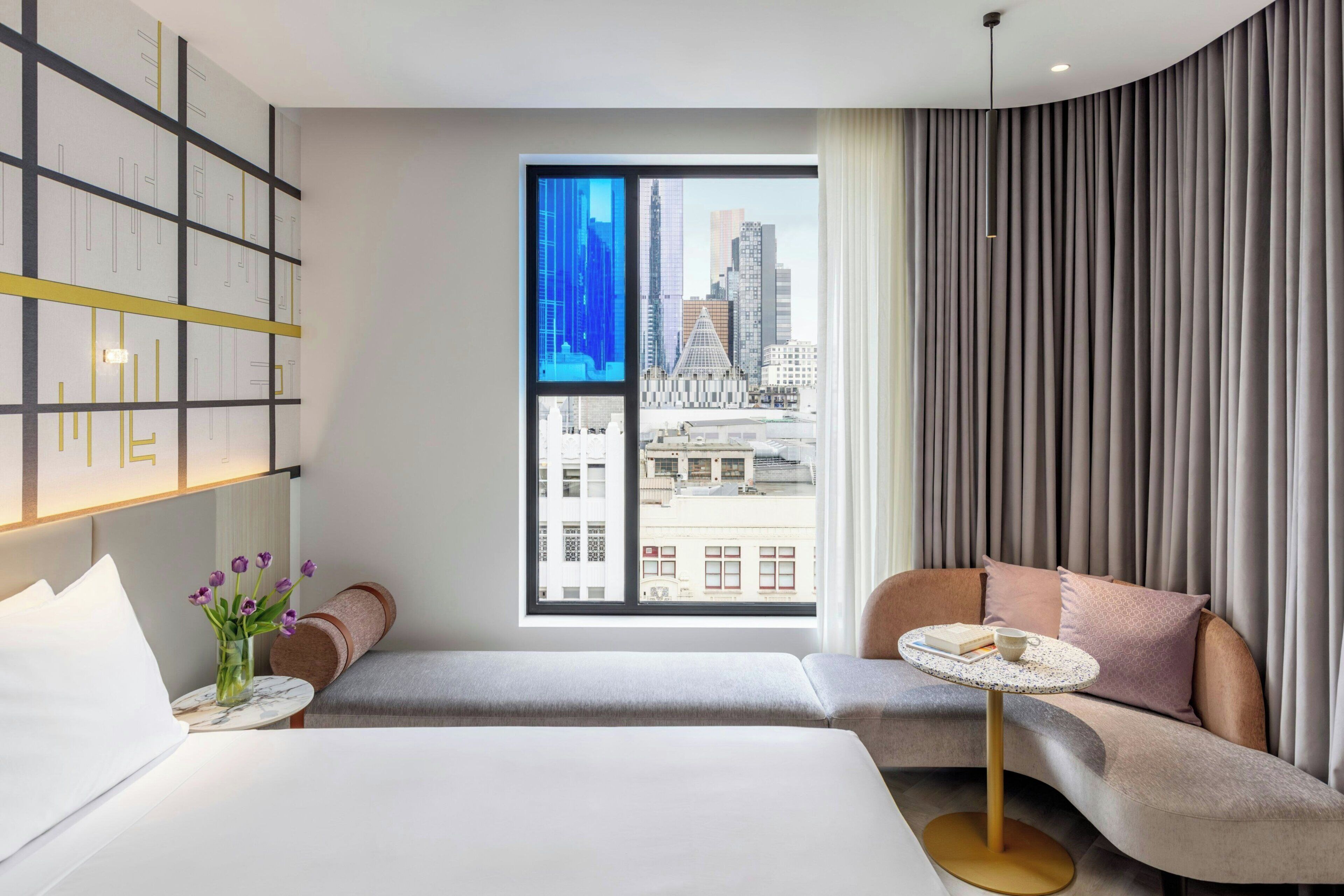 A modern hotel room with a large window, a bed, a seating area, and a bookshelf.