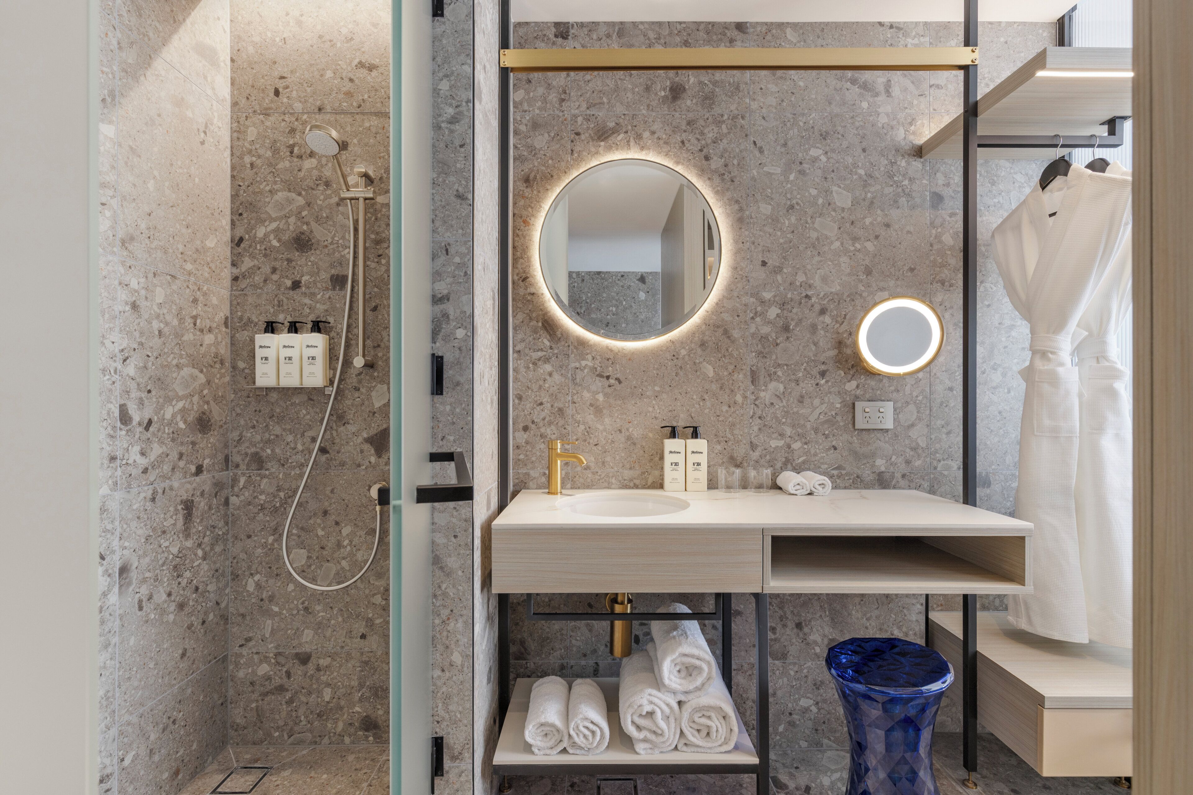 A modern bathroom with a marble backsplash, a round mirror with a light, a sink with a gold faucet, and a shower area.