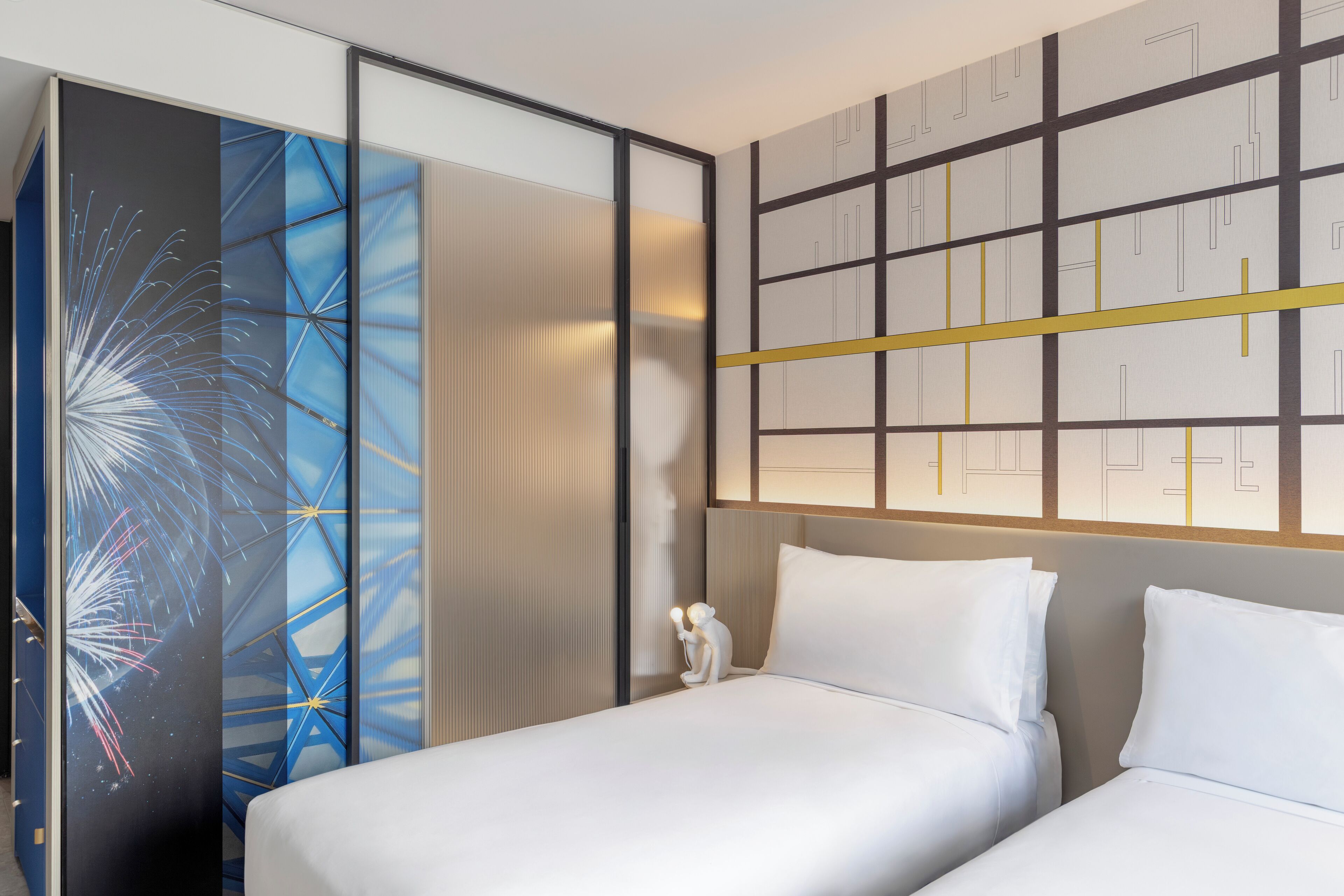 A hotel room with two beds, a decorative wall with geometric patterns, and a nightstand with a lamp.