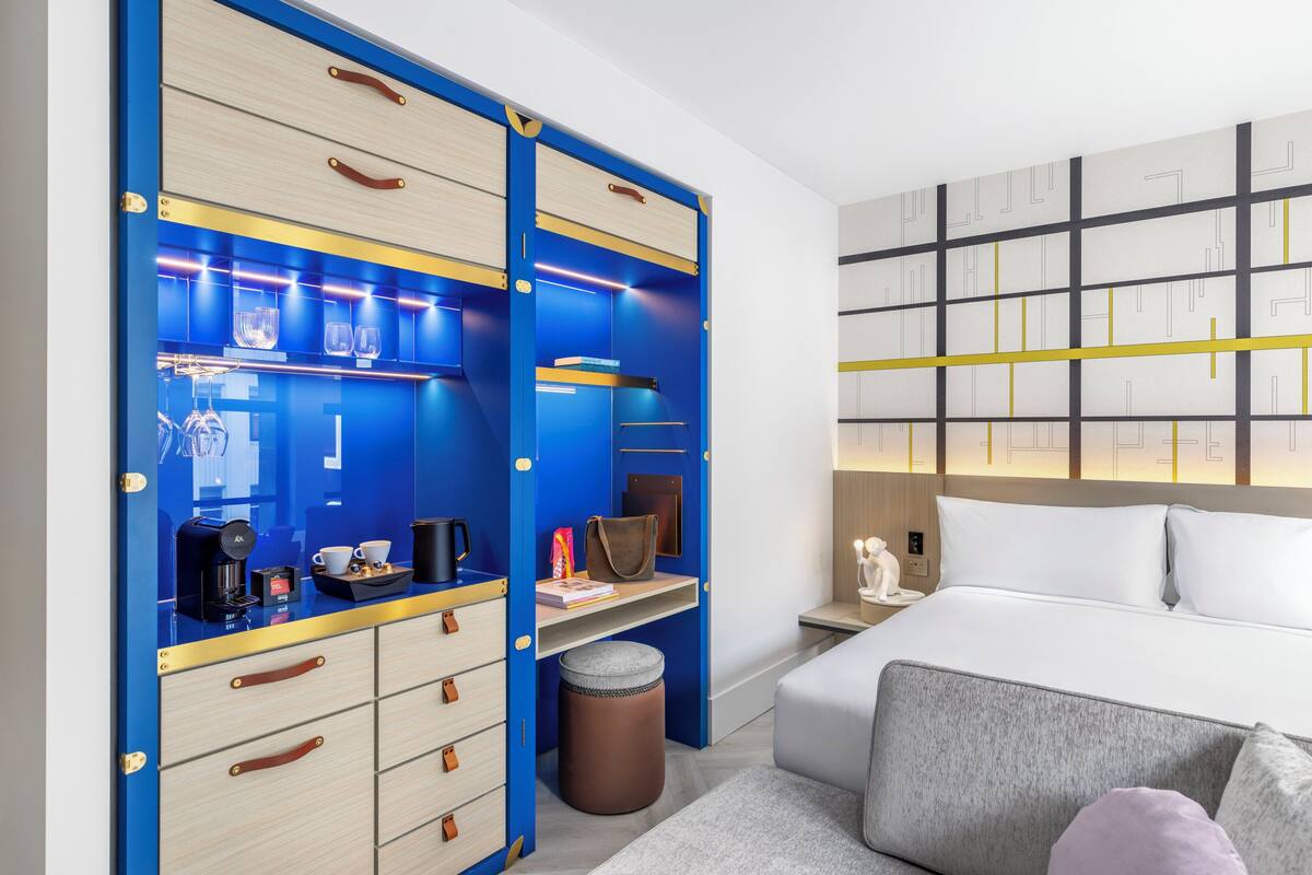 Ein modernes Hotelzimmer mit einem großen Bett, einem Schreibtisch mit Computer, einem Kleiderschrank mit Schubladen und einer Wand mit geometrischem Muster.