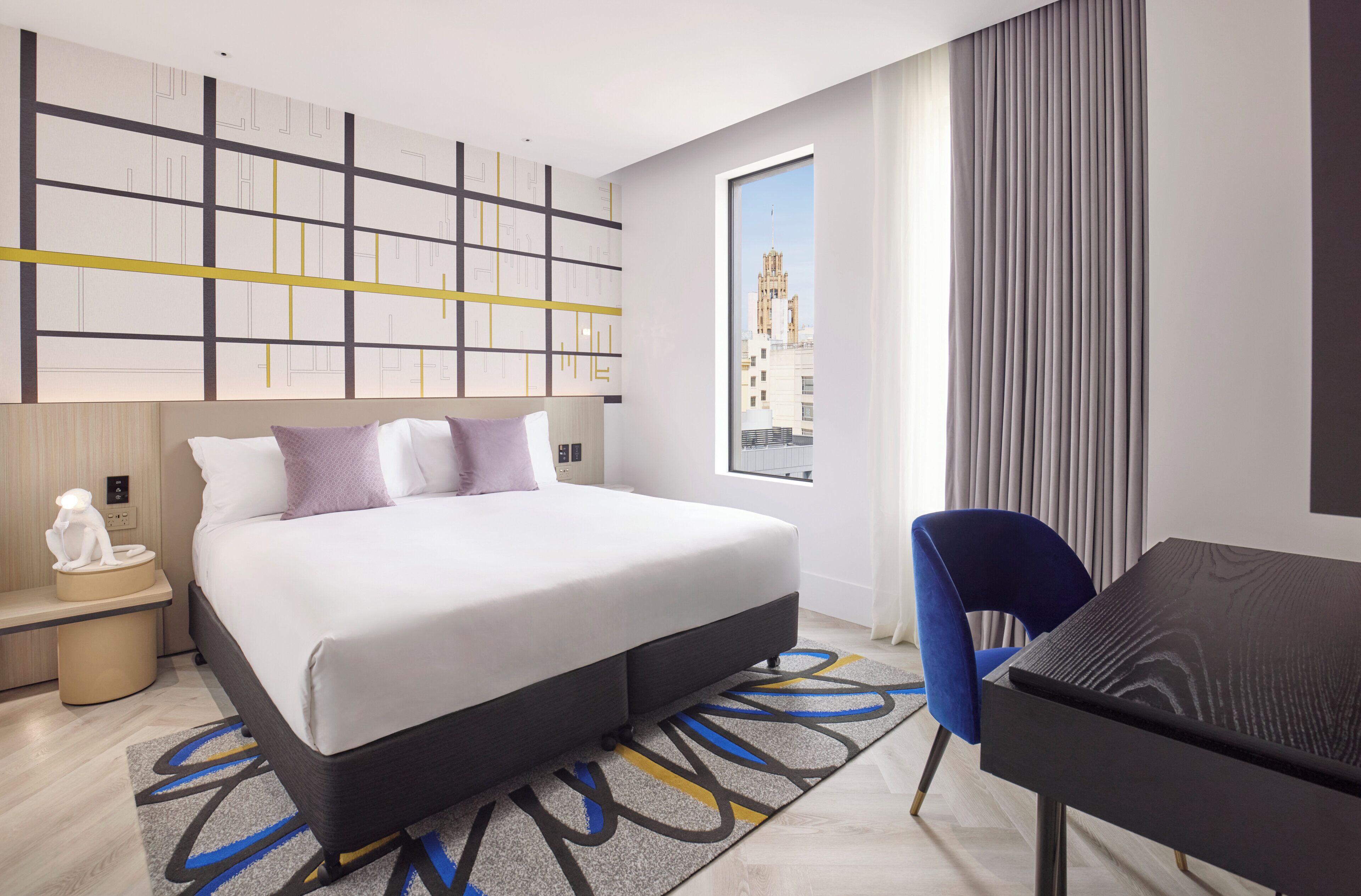 Habitación de hotel con cama, mesita de noche, lámpara, silla, escritorio y vista a un edificio a través de la ventana.