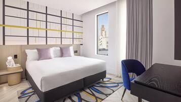 Habitación de hotel con cama, mesita de noche, lámpara, silla, escritorio y vista a un edificio a través de la ventana.