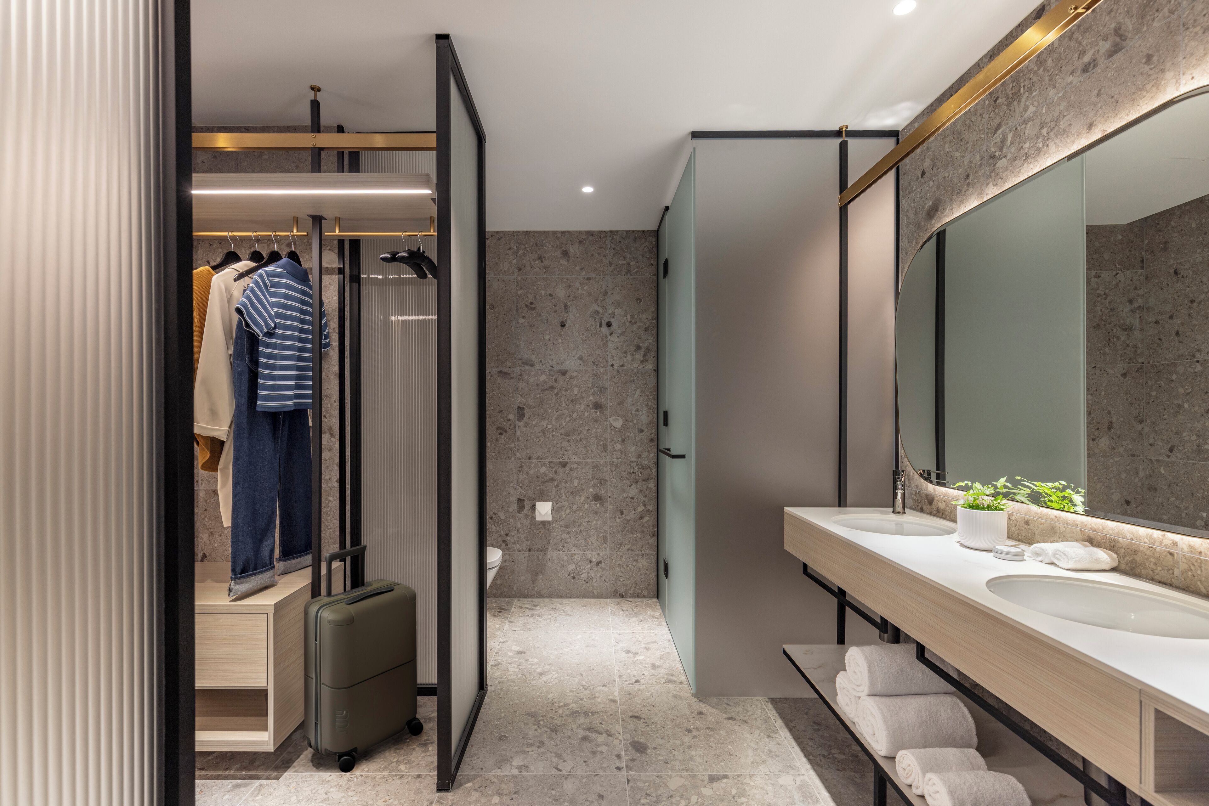 A modern bathroom with a large mirror, two sinks, a shower area, and a walk-in closet with hanging clothes and a suitcase.