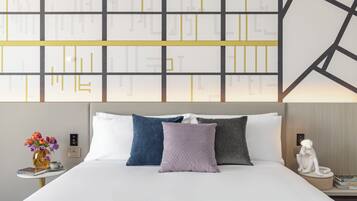 A hotel room with a large bed, multiple pillows, a nightstand with a lamp, a vase with flowers, and a wall with geometric patterns.