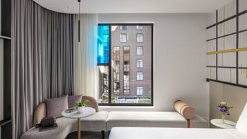A modern hotel room with a curved wall, a large window with a city view, a sofa, a small table with a plant, and a bed.