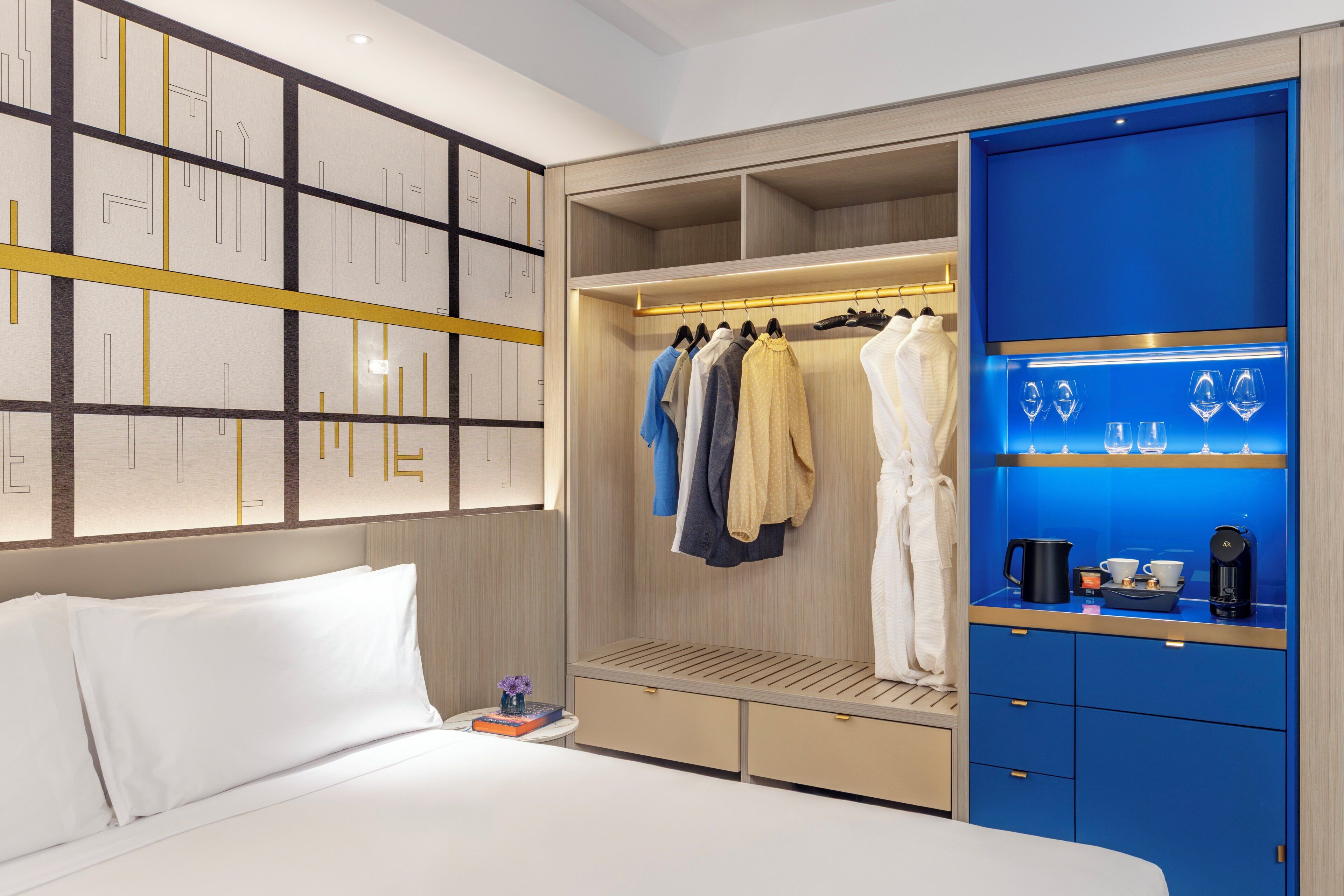 A hotel room with a bed, a wardrobe with hanging clothes, and a shelf with glasses and a coffee maker.
