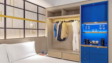 A hotel room with a bed, a wardrobe with hanging clothes, and a shelf with glasses and a coffee maker.