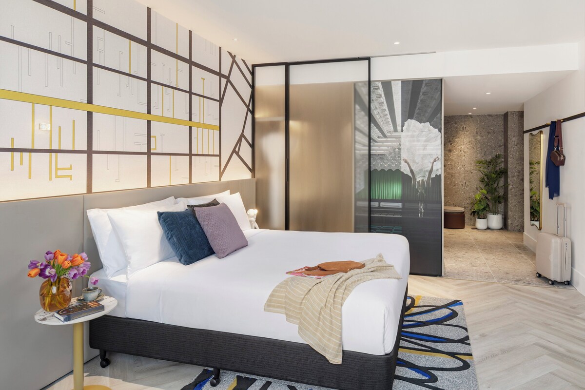 Ein modernes Hotelzimmer mit einem großen Bett, einem Nachttisch mit Blumen in einer Vase, einer Wand mit geometrischen Mustern und Blick auf die Stadt durch eine Glastür.